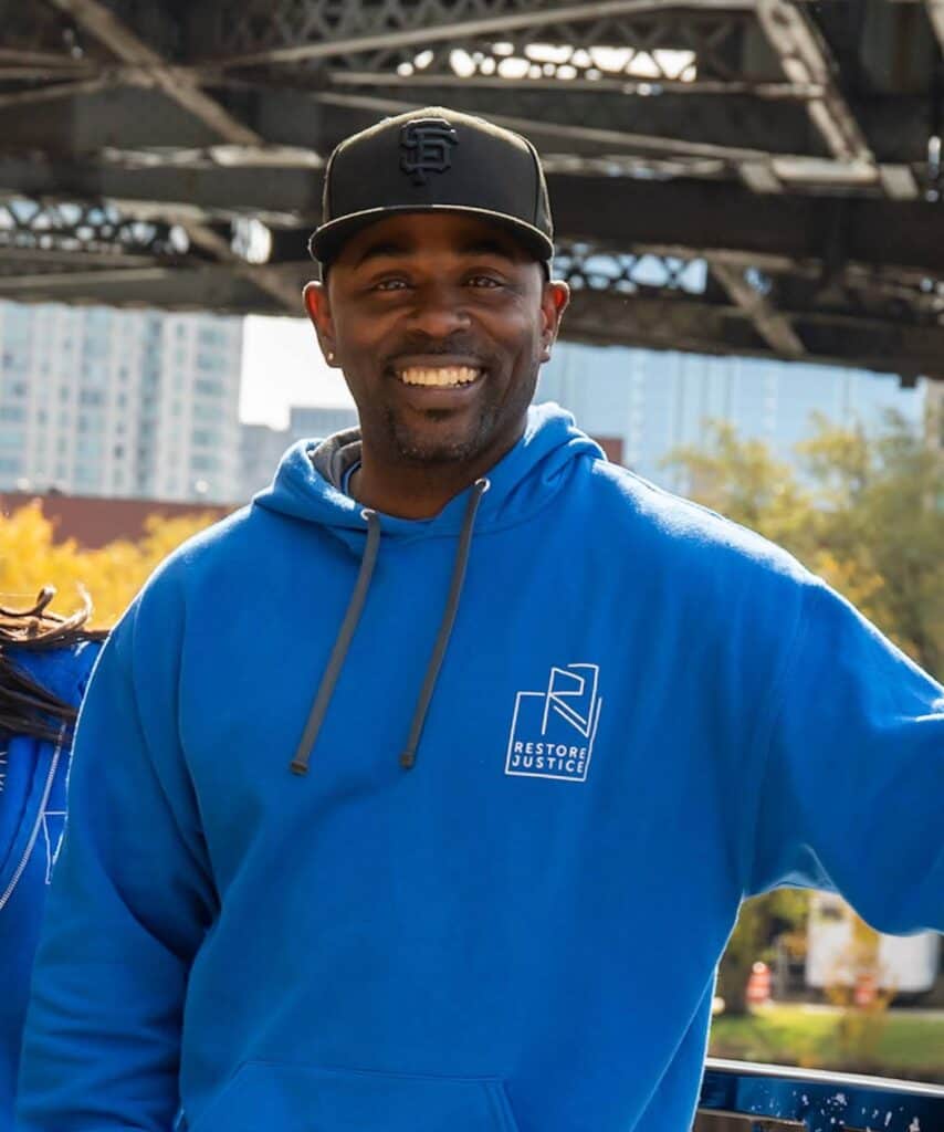 Black man smiling widely, wearing restore justice hoodie and cap, outdoors in front of a bridge