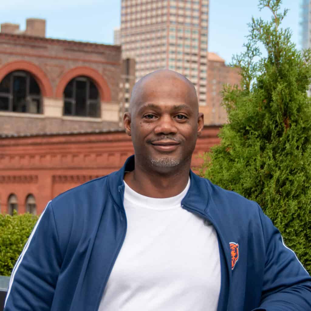 Adult black man with shaved head smiling subtly, outside in front of greenery and buildings